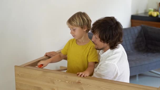 Πατέρας και γιος συναρμολογούν ξύλινα έπιπλα από μικρά μέρη. Μικρό αγόρι βοηθά τον πατέρα του για τη συναρμολόγηση ενός πίνακα. — Αρχείο Βίντεο