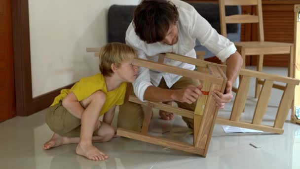 Padre e hijo montan muebles de madera de pequeñas piezas. Pequeño niño ayuda a su padre a montar una silla . — Vídeo de stock