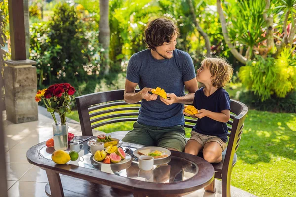 Familia de dos comiendo bien servido desayuno fuera guapo joven verter un poco de café y su lindo hijo comer deliciosa fruta en el desayuno —  Fotos de Stock