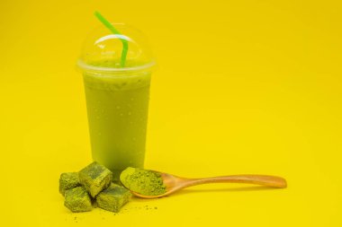 Bir plastik kap ve saman ve tozu matcha sarı zemin üzerine kaşıkla buzlu yeşil çay latte. Ev yapımı Matcha Latte çayları ile süt sıfır atık