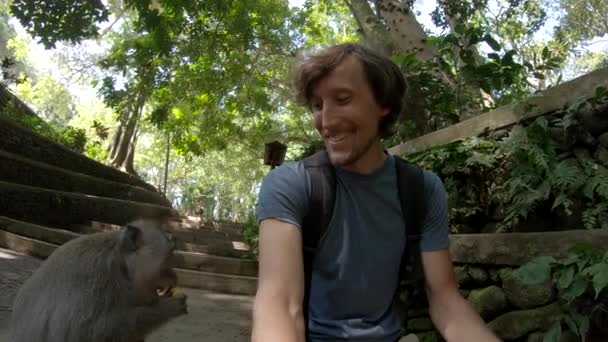 Filmagem em câmera lenta de um jovem alimentando e fazendo uma selfie com um macaco macaco no parque natural da floresta de macacos na aldeia de Ubud, Bali — Vídeo de Stock