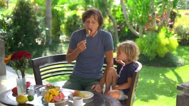 Медленный снимок молодого отца и его маленького сына, завтракающего тостами с авокадо и большим количеством фруктов на террасе в окружении сада — стоковое видео