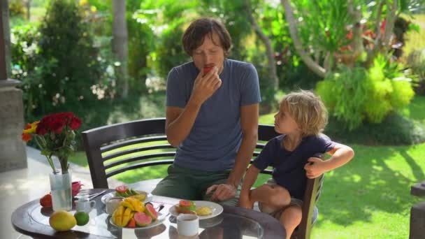 Fotografía en cámara lenta de un joven padre y su pequeño hijo desayunando tostadas de aguacate y muchas frutas en su terraza rodeados de un jardín — Vídeos de Stock