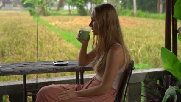 Slowmotion tiro de uma jovem mulher bebendo chá verde matcha latte em um café tropical com vista para um campo de arroz — Vídeo de Stock