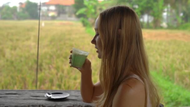 Slowmotion πλάνο του μια νεαρή γυναίκα που πίνει latte matcha πράσινο τσάι σε μια τροπική καφετέρια με θέα σε έναν ορυζώνα — Αρχείο Βίντεο