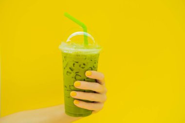Yeşil çay sütlü buzun içinde plastik kap ve saman sarı zemin üzerine sarı manikür ile kadın elinde. Ev yapımı buzlu Matcha Latte sütlü çay götürün