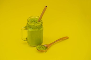 Mason kavanoza ve saman ve tozu matcha sarı zemin üzerine kaşıkla buzlu yeşil çay latte. Ev yapımı Matcha Latte çayları ile süt sıfır atık