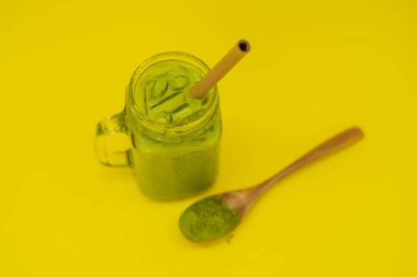 Mason kavanoza ve saman ve tozu matcha sarı zemin üzerine kaşıkla buzlu yeşil çay latte. Ev yapımı Matcha Latte çayları ile süt sıfır atık