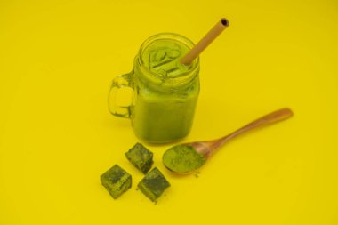 Mason kavanoza ve saman buzlu yeşil çay kahve ve kaşık toz matcha ve şeker ile matcha sarı zemin üzerine yapılmış. Ev yapımı Matcha Latte çayları ile süt sıfır atık