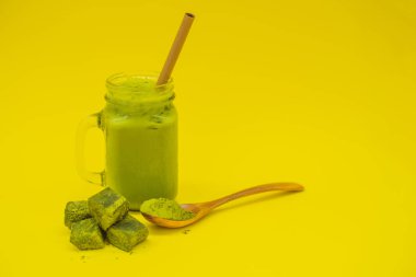 Mason kavanoza ve saman buzlu yeşil çay kahve ve kaşık toz matcha ve şeker ile matcha sarı zemin üzerine yapılmış. Ev yapımı Matcha Latte çayları ile süt sıfır atık