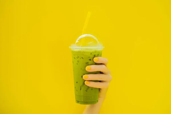 Grüntee-Latte mit Eis in Plastikbecher und Stroh in weiblicher Hand mit gelber Maniküre auf gelbem Hintergrund. hausgemachter Eistee Matcha Latte Tea mit Milch zum Mitnehmen — Stockfoto