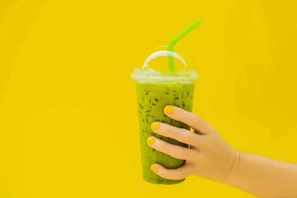 Grüntee-Latte mit Eis in Plastikbecher und Stroh in weiblicher Hand mit gelber Maniküre auf gelbem Hintergrund. hausgemachter Eistee Matcha Latte Tea mit Milch zum Mitnehmen — Stockfoto