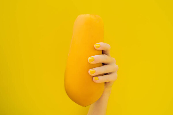 Hand with a yellow manicure holding a orange ripe papaya on a yellow background