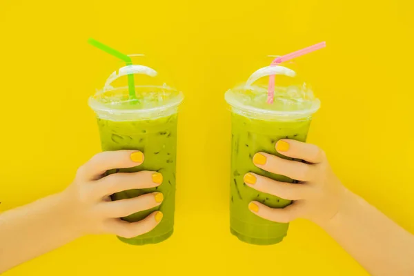 Zwei Grüntee-Latte mit Eis in Plastikbecher und Stroh in weiblicher Hand mit gelber Maniküre auf gelbem Hintergrund. hausgemachter Eistee Matcha Latte Tea mit Milch zum Mitnehmen — Stockfoto