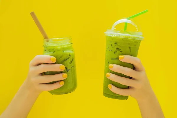Зеленый чай латте со льдом в банке каменщика и в пластиковой чашке в руках на желтом фоне. Домашний чай маття с молоком без отходов — стоковое фото