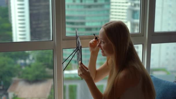 Super Slow Motion toma de una joven haciendo su maquillaje sittng por una ventana panorámica en el centro de una ciudad con rascacielos en un segundo plano — Vídeo de stock