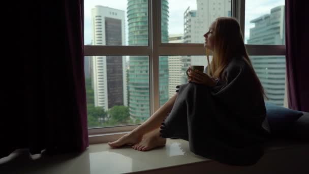 Foto de Super Slow Motion de una joven que bebe té o café envuelta en una tela a cuadros sentada junto a una ventana panorámica en el centro de una ciudad con rascacielos en un segundo plano — Vídeos de Stock