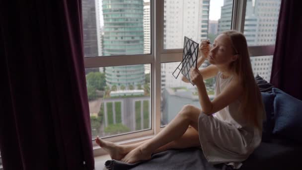 Filme Super Slow Motion de uma jovem mulher fazendo sua maquiagem sentada por uma janela panorâmica no centro da cidade com arranha-céus em um fundo — Vídeo de Stock