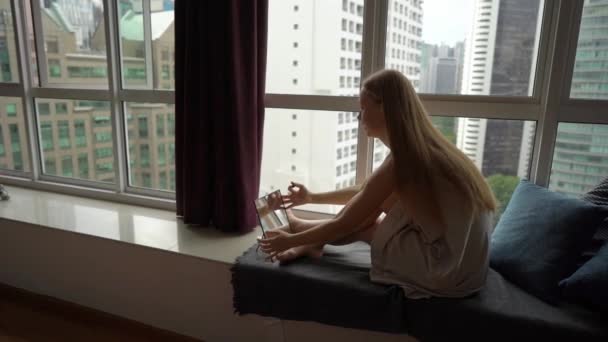 Super Slow Motion toma de una joven haciendo su maquillaje sittng por una ventana panorámica en el centro de una ciudad con rascacielos en un segundo plano — Vídeo de stock