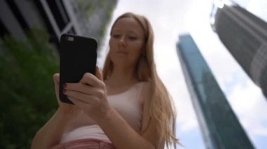 Bir genç kadın thar Superslowmotion atış gökdelenler, bir arka plan ile kent merkezinde bir akıllı telefon kullanın. Şehir kavramı arasında gezinti. Toplu taşıma kavramı. Taksi rezervasyon kavramı