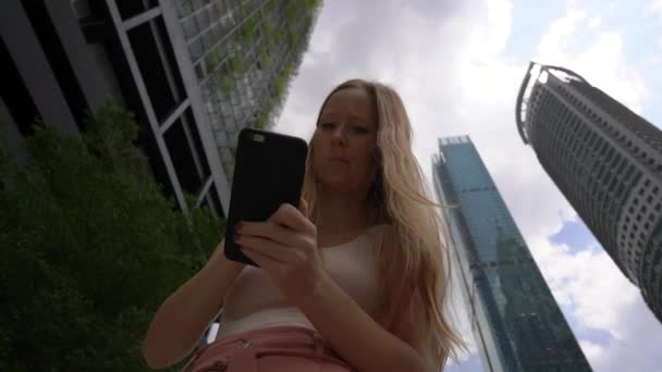 Bir genç kadın thar Superslowmotion atış gökdelenler, bir arka plan ile kent merkezinde bir akıllı telefon kullanın. Şehir kavramı arasında gezinti. Toplu taşıma kavramı. Taksi rezervasyon kavramı — Stok video