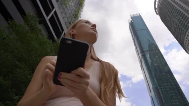 Superslowmotion πλάνο του μια νεαρή γυναίκα thar να χρησιμοποιήσετε ένα smartphone στο κέντρο της πόλης περιοχή με ουρανοξύστες στο φόντο. Πλοήγηση μέσω της έννοιας της πόλης. Έννοια των δημόσιων μεταφορών. Έννοια κράτηση ταξί — Αρχείο Βίντεο