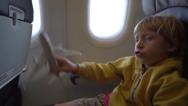 Slowmotion shot of a little boy that plays with white toy airplane sitting in a chair onboard of an airplane. Freedom concept. Childhood concept. Children travel concept — Stock Video