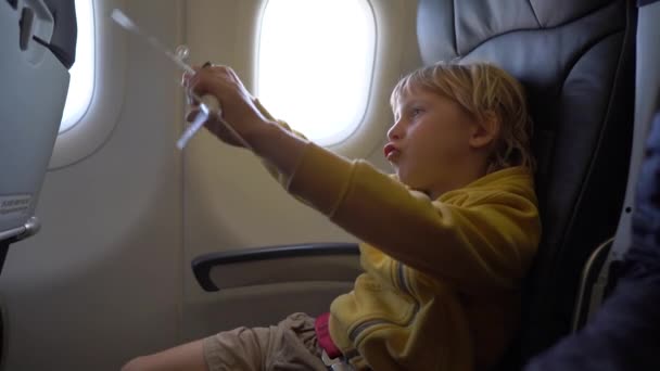 Filmagem em câmera lenta de um menino que brinca com um avião de brinquedo branco sentado em uma cadeira a bordo de um avião. Conceito de liberdade. Conceito de infância. Conceito de viagem infantil — Vídeo de Stock