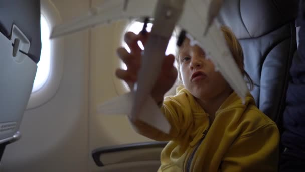 Filmagem em câmera lenta de um menino que brinca com um avião de brinquedo branco sentado em uma cadeira a bordo de um avião. Conceito de liberdade. Conceito de infância. Conceito de viagem infantil — Vídeo de Stock