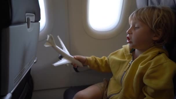 Filmagem em câmera lenta de um menino que brinca com um avião de brinquedo branco sentado em uma cadeira a bordo de um avião. Conceito de liberdade. Conceito de infância. Conceito de viagem infantil — Vídeo de Stock
