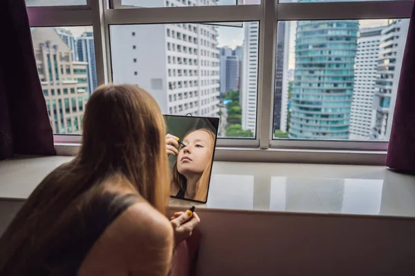 Молодая женщина делает макияж сидя у окна с панорамным видом на небоскребы и большой город — стоковое фото