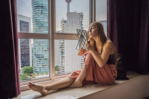 年轻女子坐在窗边化妆, 可以看到摩天大楼和大城市的全景 — 图库照片