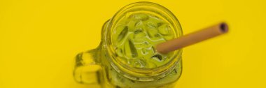 Buz mason kavanoza ve saman sarı zemin üzerine yeşil çay kahve. Ev yapımı buzlu Matcha Latte sütlü çay sıfır atık afiş, uzun biçimde