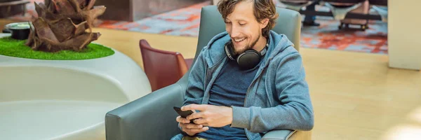 Мужчина в зале ожидания аэропорта ждет своего самолета, пользуется смартфоном и наушниками. Молодой улыбающийся человек с бородой держит смартфон в руках, работая в аэропорту, ожидая самолет — стоковое фото