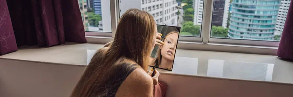 Mujer joven hace maquillaje sentado junto a la ventana con una vista panorámica de los rascacielos y la gran ciudad BANNER, FORMATO LARGO — Foto de Stock