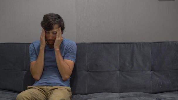 A young man sits on a couch and man feels upset and confused. Copy space on the right side — Stock Video
