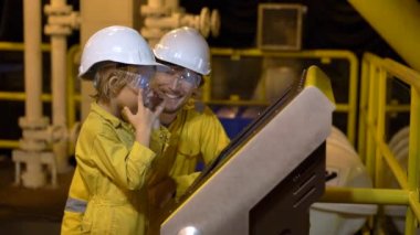 Genç bir adam ve küçük bir çocuk sarı iş üniformalı, gözlük, her ikisi de ve endüstriyel bir ortamda kask, Platform veya sıvılaştırılmış gaz tesisi bir ekrana bakarak petrol. Slowmotion atış