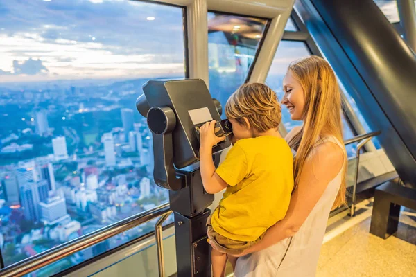 Mom and son are looking at Kuala lumpur cityscape. Use binoculars. Panoramic view of Kuala Lumpur city skyline evening at sunset skyscrapers building in Malaysia. Traveling with kids concept — Stock Photo, Image