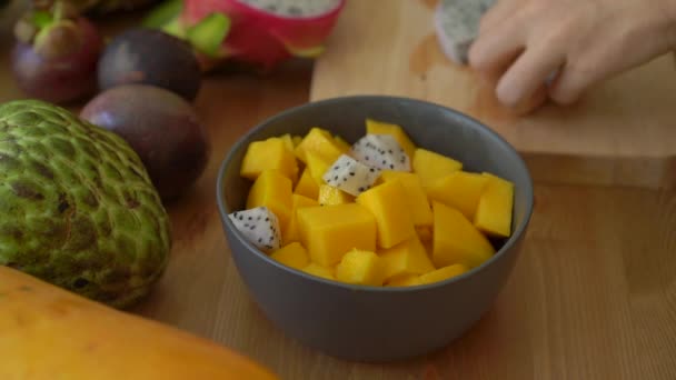 2x vezes Slowmotion tiro de uma jovem mulher colocando os cubos de frutas do dragão em uma tigela de cerâmica cinza e muitas frutas tropicais estavam em uma mesa — Vídeo de Stock