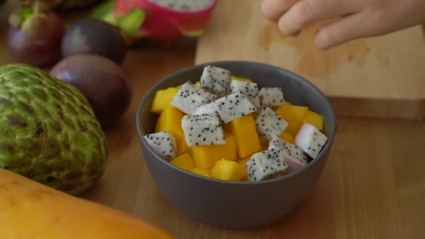 2x vezes Slowmotion tiro de uma jovem mulher colocando os cubos de frutas do dragão em uma tigela de cerâmica cinza e muitas frutas tropicais estavam em uma mesa — Vídeo de Stock