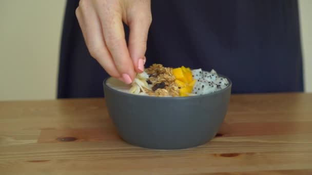 Dragon meyve, mango, granola, Kuru üzüm, badem dilimleri ve chia tohum ile bir genç kadın pişirme muz smoothie kase kadeh Slowmotion — Stok video