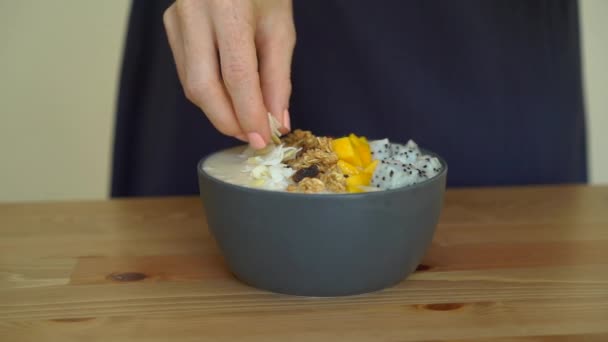 Slowmotion skott av en ung kvinna matlagning banana smoothie bowl med dragon frukt, mango, granola, russin, mandel skivor och chiafrön — Stockvideo