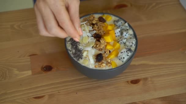 Slowmotion skott av en ung kvinna matlagning banana smoothie bowl med dragon frukt, mango, granola, russin, mandel skivor och chiafrön — Stockvideo