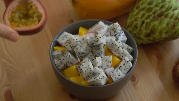 Plan lent d'une jeune femme mettant le fruit de la passion coupé dans le bol avec un fruit du dragon et des cubes de manfo et beaucoup de fruits tropicaux sur une table — Video