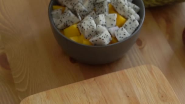 Slowmotion shot of a young woman putting the cut passion fruit into the bowl with a dragon fruit and manfo cubes and lots of tropical fruits lay on a table — Stock Video