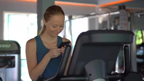 Uma jovem mulher no ginásio em uma bicicleta de exercício segurando o telefone na frente de seus olhos em suas mãos. Toda a atenção está no telefone. O conceito de dependência das redes sociais. Dependência móvel — Vídeo de Stock