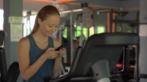 Молода жінка в спортзалі на тренажерному велосипеді тримає телефон перед очима в руках. Вся увага в телефоні. Поняття залежності від соціальних мереж. Мобільна залежність — стокове відео
