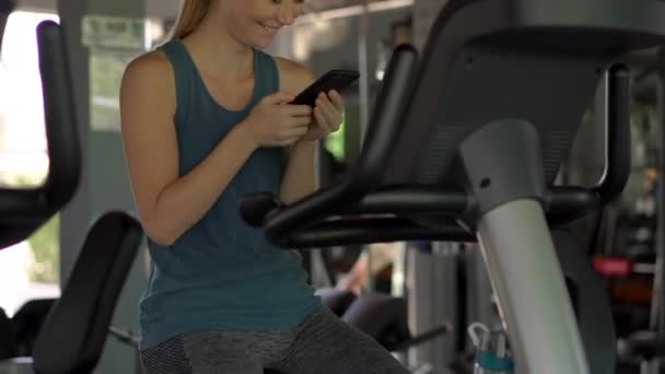 Uma jovem mulher no ginásio em uma bicicleta de exercício segurando o telefone na frente de seus olhos em suas mãos. Toda a atenção está no telefone. O conceito de dependência das redes sociais. Dependência móvel — Vídeo de Stock