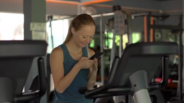 En ung kvinna på gymmet på en motionscykel som håller telefonen framför hennes ögon i hennes händer. All uppmärksamhet är i telefonen. Begreppet beroende på sociala nätverk. Mobila missbruk — Stockvideo