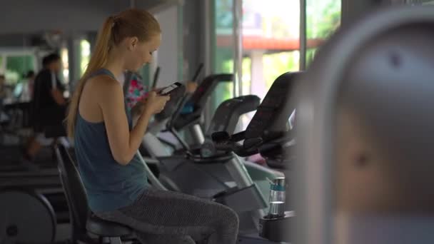 Uma jovem mulher no ginásio em uma bicicleta de exercício segurando o telefone na frente de seus olhos em suas mãos. Toda a atenção está no telefone. O conceito de dependência das redes sociais. Dependência móvel — Vídeo de Stock